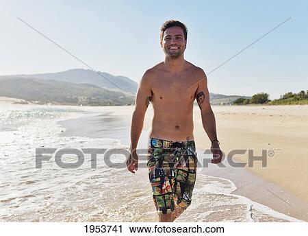 Stock Image - a shirtless man walking down the beach in the tide; tarifa cadiz andalusia spain. Fotosearch