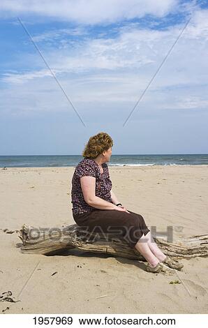 Stock Photo - a woman sits on a piece of driftwood on the beach along the coast; northumberland england. Fotosearch