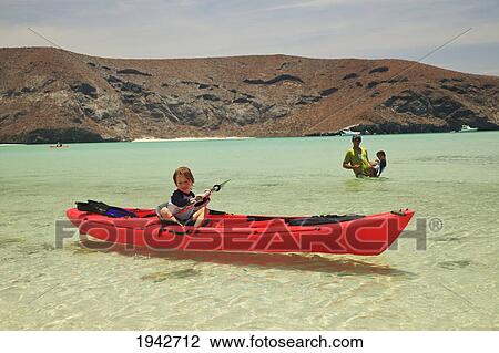 Stock Image - A Young Boy Sits In A Red Boat By The Shore While A Father And Son Play Out In The Water At Los Islotes National Marine Park Espiritu Santo Island; La Paz Baja California Mexico. Fotosearch