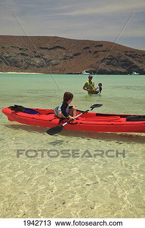 Stock Image - A Young Boy Sits In A Red Boat By The Shore While A Father And Son Play Out In The Water At Los Islotes National Marine Park Espiritu Santo Island; La Paz Baja California Mexico. Fotosearch