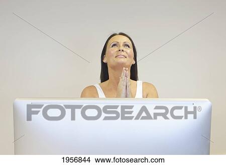 Picture - a young woman looks upwards with praying hands in front of her computer screen; south shields tyne and wear england. Fotosearch