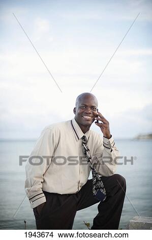 Picture - Businessman Using A Cell Phone At The Water's Edge; South Caicos Turks And Caicos Islands. Fotosearch