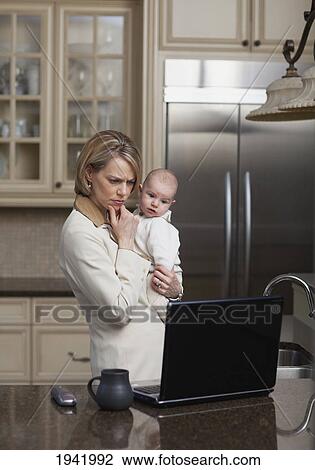 Stock Image - Businesswoman Working On Computer At Home And Holding Baby; Jordan Ontario Canada. Fotosearch