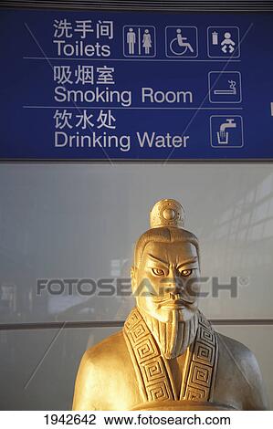 Stock Image - Chinese Statue Under A Sign In The Beijing Airport With Translation In Chinese And English; Beijing China. Fotosearch