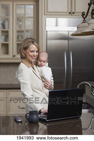 Smiling Businesswoman Working On Computer At Home And Holding Baby; Jordan Ontario Canada View Large Photo Image Stock Image - Smiling Businesswoman Working On Computer At Home And Holding Baby; Jordan Ontario Canada. Fotosearch