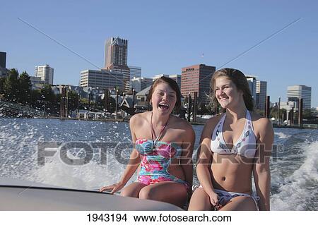Picture - Two Teenage Girls Sitting On The Back Of A Boat Wearing Bathing Suits With The City In The Background; Portland Oregon United States Of America. Fotosearch