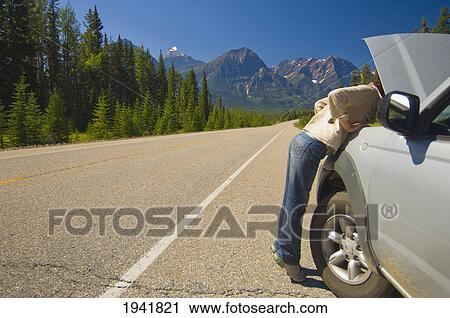 Stock Image - Woman Looking Under Hood Of Her Car On Mountain Road; Alberta Canada. Fotosearch