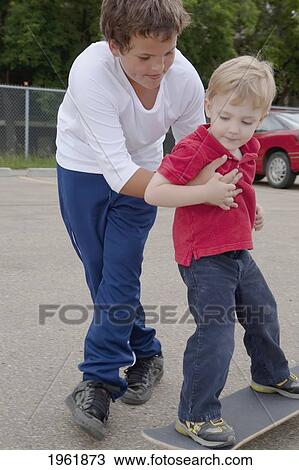 Stock Image - a boy teaches a younger boy how to skateboard; edmonton, alberta, canada. Fotosearch