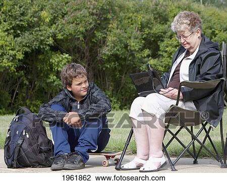 Stock Image - a senior woman sits outside using a laptop computer while a boy watches her; edmonton, alberta, canada. Fotosearch