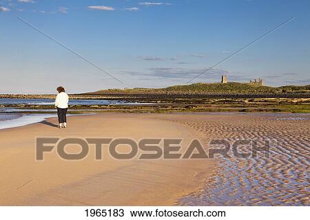 Stock Image - a woman stands on the beach looking out over the ocean with dunstanburgh castle in the background; northumberland england. Fotosearch