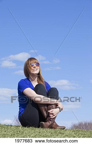 Stock Image - a young woman wearing sunglasses and sitting on the grass; edmonton alberta canada. Fotosearch