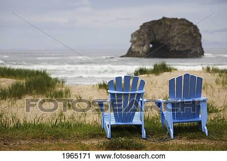 Stock Image - two blue adirondack chairs on a grassy beach with rock formations in the ocean; rockaway beach oregon united states of america. Fotosearch