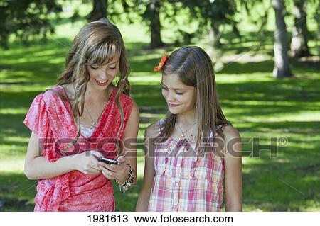 Stock Image - two sisters together in a park looking at a cell phone; edmonton alberta canada. Fotosearch
