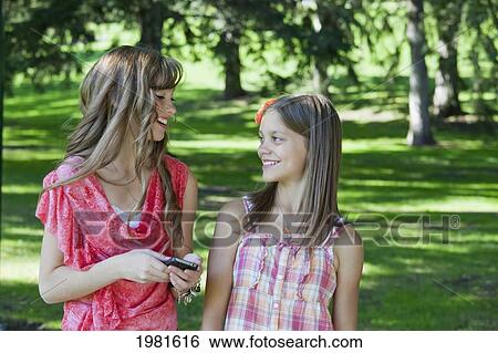 Stock Photograph - two sisters together in a park looking at a cell phone; edmonton alberta canada. Fotosearch