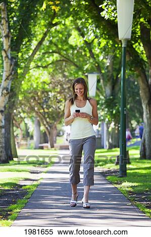 Stock Photography - woman walking down a park path and checking text messages; edmonton alberta canada. Fotosearch