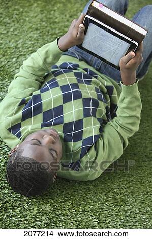 Picture - A boy in an argyle sweater lays on the grass reading from an electronic notebook; portland oregon united states of america. Fotosearch