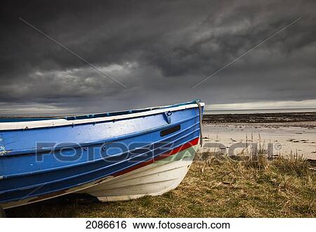 Stock Photograph - A colourful rowboat on the shore with dark storm clouds overhead; boulmer northumberland england. Fotosearch