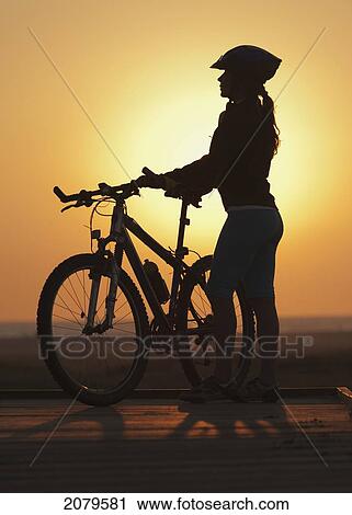 Stock Image - A cyclist stands on the beach with her bike at sunset; tarifa cadiz andalusia spain. Fotosearch