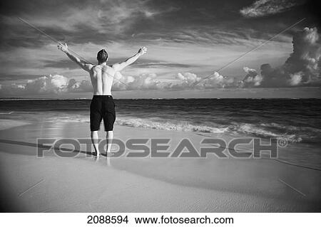 Picture - A man stands on a wet beach with arms raised looking out at the ocean; punta cana la altagracia dominican republic. Fotosearch