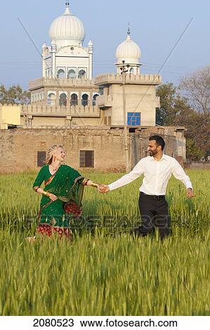 Stock Image - A mixed race couple her wearing a sari holding hands and running through a field with a temple in the background; ludhiana punjab india. Fotosearch