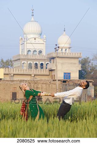 Stock Photography - A mixed race couple her wearing a sari holding hands and dancing in a field with a temple in the background; ludhiana punjab india. Fotosearch