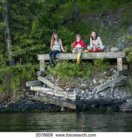 Stock Photo - A mother and two daughters sit on a wooden dock at the edge of a lake; lake of the woods ontario canada. Fotosearch