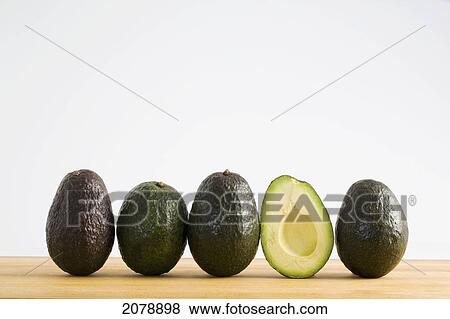 Stock Photo - A row of avocados standing upright on a wooden board with one cut in half without the pit; calgary alberta canada. Fotosearch