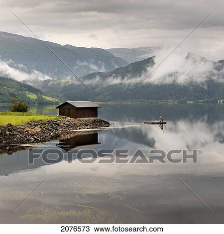 Stock Image - A small wooden building at the water's edge and a dock in a tranquil lake; granvin hordaland norway. Fotosearch