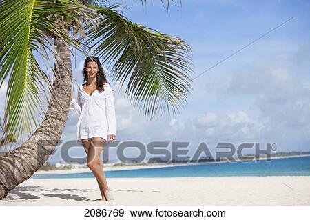 Stock Photo - A woman in a white dress posing beside a palm tree on a beach; punta cana la altagracia dominican republic. Fotosearch