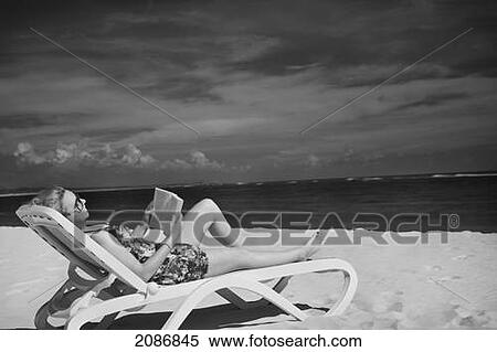 Stock Photography - A woman sits reading in a lounge chair on the beach; punta cana la altagracia dominican republic. Fotosearch