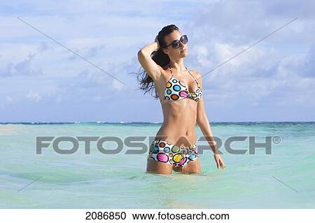 Stock Image - A woman standing in the ocean wearing a colourful bikini and sunglasses; punta cana la altagracia dominican republic. Fotosearch