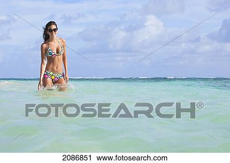Stock Image - A woman standing in the ocean wearing a colourful bikini and sunglasses; punta cana la altagracia dominican republic. Fotosearch