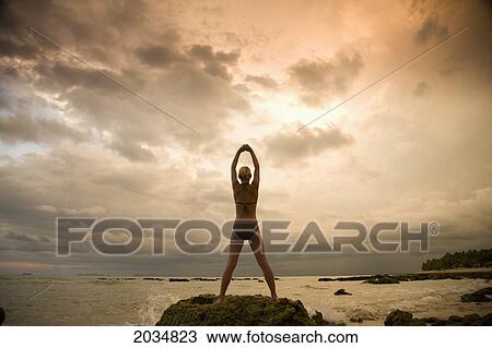 Stock Image - A Woman Tourist Wearing A Bikini Raises Her Arms And Does Yoga On The Beach Of A Tropical Island At Sunset; Koh Lanta Thailand. Fotosearch