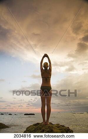 Stock Photograph - A Woman Tourist Wearing A Bikini Raises Her Arms And Does Yoga On The Beach Of A Tropical Island At Sunset; Koh Lanta Thailand. Fotosearch