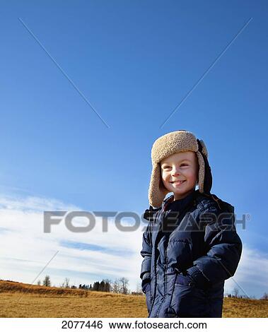 Stock Photograph - A young boys stands on a grass field wearing a coat and hat; spruce grove alberta canada. Fotosearch