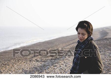 Stock Image - A young woman stands on the rocky beach at the water's edge; aldeburgh england. Fotosearch