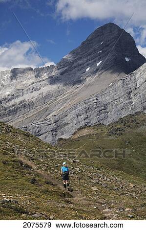 Stock Photo - Female Hiker In The Distance On A Mountain Trail With Mountain Peak In The Background And Blue Sky With Clouds; Alberta Canada. Fotosearch