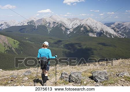 Stock Image - Female Hiker With Hiking Poles Along Ridge Trail With Mountians And Valley In The Distance And Blue Sky And Clouds; Alberta Canada. Fotosearch