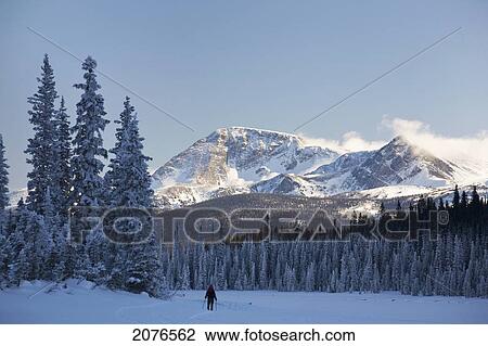 Stock Image - Female snowshoeing on snow covered lake with snow covered evergreens in the shade mountains and clouds rolling on peaks in sunlight and blue sky; lake louise alberta canada. Fotosearch