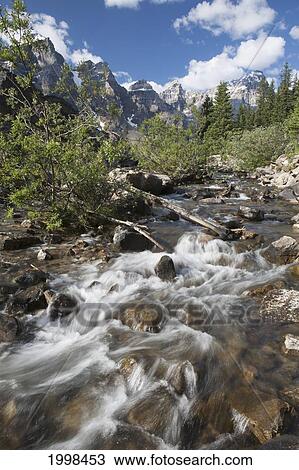 Flowing Stream With Shrubs Along It's Banks With Mountains In The Distance And Blue Sky And Clouds; Alberta Canada View Large Photo Image Stock Image - Flowing Stream With Shrubs Along It's Banks With Mountains In The Distance And Blue Sky And Clouds; Alberta Canada. Fotosearch
