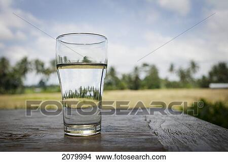 Picture - Glass of water on a table with rice fields in the background; bali indonesia. Fotosearch