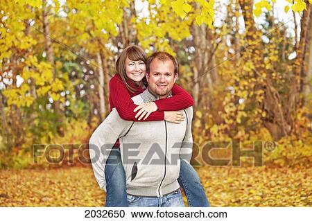 Stock Image - Husband Carrying His Wife On His Back In A Park In Autumn; Edmonton Alberta Canada. Fotosearch