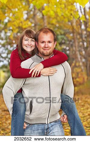Stock Image - Husband Carrying His Wife On His Back In A Park In Autumn; Edmonton Alberta Canada. Fotosearch