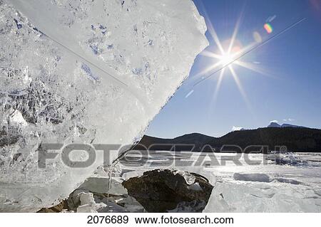Stock Photo - Ice formation on shoreline of a mountain lake with a sunburst and blue sky; banff alberta canada. Fotosearch