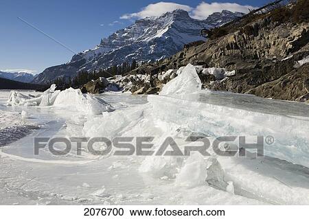 Stock Image - Ice formations on a frozen lake shoreline with mountains in the background and blue sky; alberta canada. Fotosearch