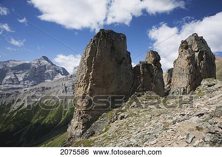 Stock Photograph - Large Rock Sentinels On Mountain Ridge With A Mountain Range And Valley In The Background With Blue Sky And Clouds; Alberta Canada. Fotosearch