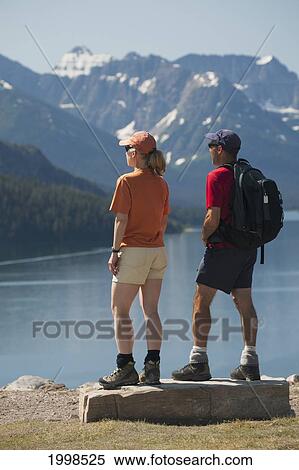 Stock Photography - Male And Female Hikers Standing On A Rock Looking Out Onto A Lake With Mountains And Blue Sky; Waterton Alberta Canada. Fotosearch