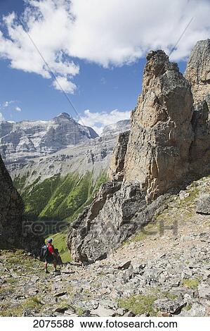 Stock Photo - Male Hiker In The Distance On Rocky Trail With Large Sentinels And Mountains In The Background With Blue Sky And Clouds; Alberta Canada. Fotosearch