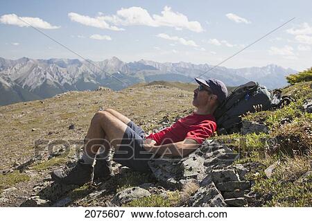 Male Hiker Laying In Meadow With Mountain Range In The Distance And Blue Sky And Clouds; Alberta Canada View Large Photo Image Stock Photo - Male Hiker Laying In Meadow With Mountain Range In The Distance And Blue Sky And Clouds; Alberta Canada. Fotosearch
