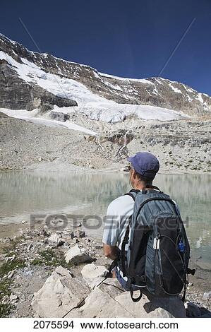 Picture - Male Hiker Sitting On A Rock With Glacier On Mountain Side Streaming Down Into A Reflective Mountain Pond With Blue Sky; Field British Columbia Canada. Fotosearch